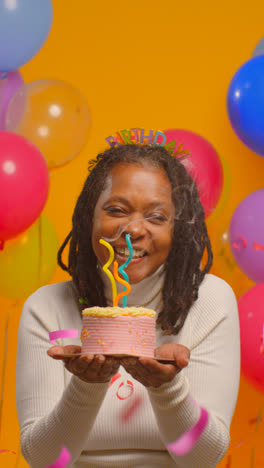 Vertical-Video-Studio-Portrait-Of-Woman-Wearing-Birthday-Headband-Celebrating-Birthday-Blowing-Out-Candles-On-Cake-With-Confetti